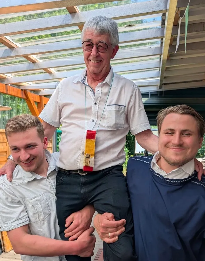 Ein älterer Mann mit Brille und weißem Haar steht lächelnd zwischen zwei jüngeren Männern. Der Mann in der Mitte trägt ein hellblaues Hemd, während die beiden anderen jeweils ein gemustertes Hemd und eine marineblaue Jacke tragen.