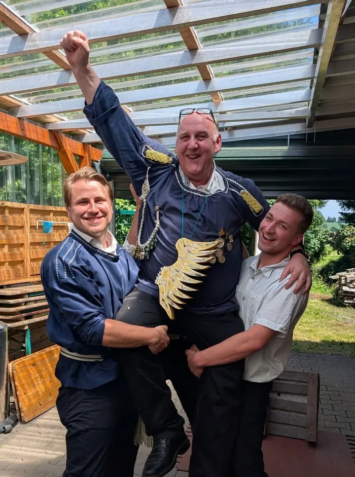 Drei Männer posieren lächelnd für ein Foto. Ein Mann in traditioneller Tracht mit einer goldenen Verzierung auf der Brust wird von zwei anderen Männern getragen.