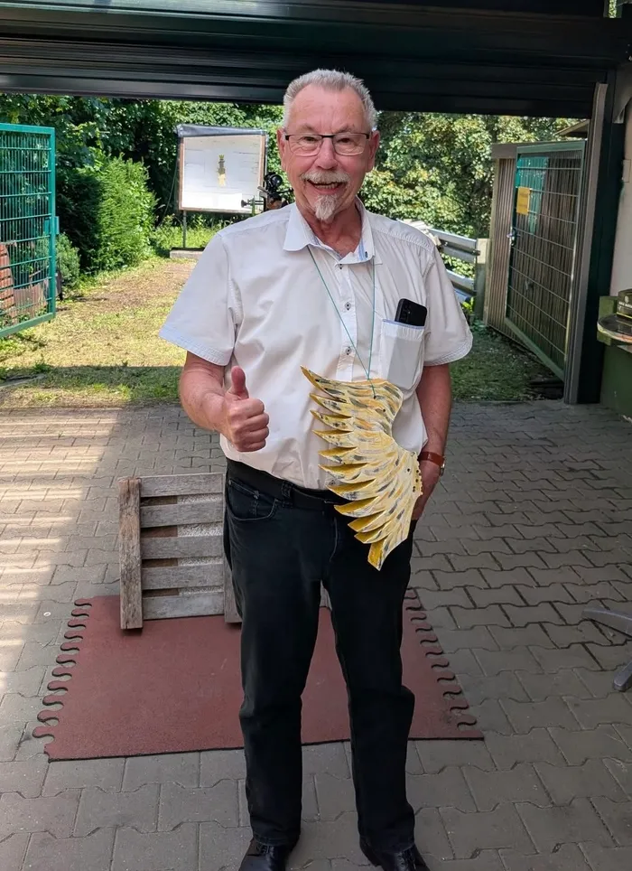 Ein älterer Mann mit Brille, weißem Haar und Bart lächelt in die Kamera und zeigt den Daumen nach oben. Er hält eine große, gelbe Blüte aus Papier oder Stoff in der Hand und steht vor einer Garage mit einem gepflasterten Hof.