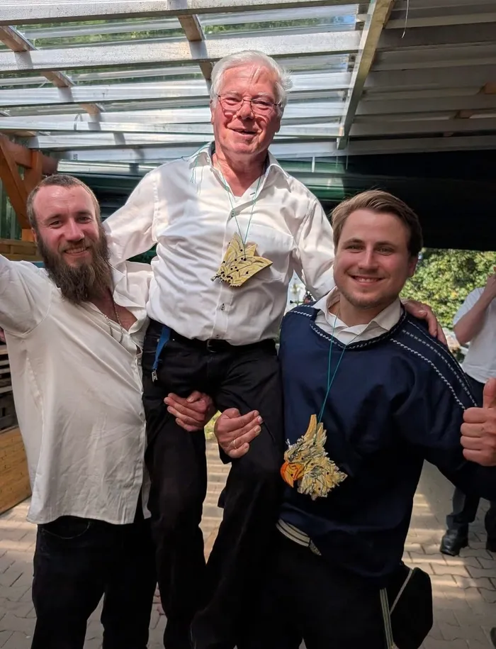 Drei Männer posieren für ein Foto. Der Mann in der Mitte, Hans Pistor, trägt ein weißes Hemd und eine Kette mit einem Wappen. Links und rechts von ihm stehen jüngere Männer, die ebenfalls Wappenketten tragen.