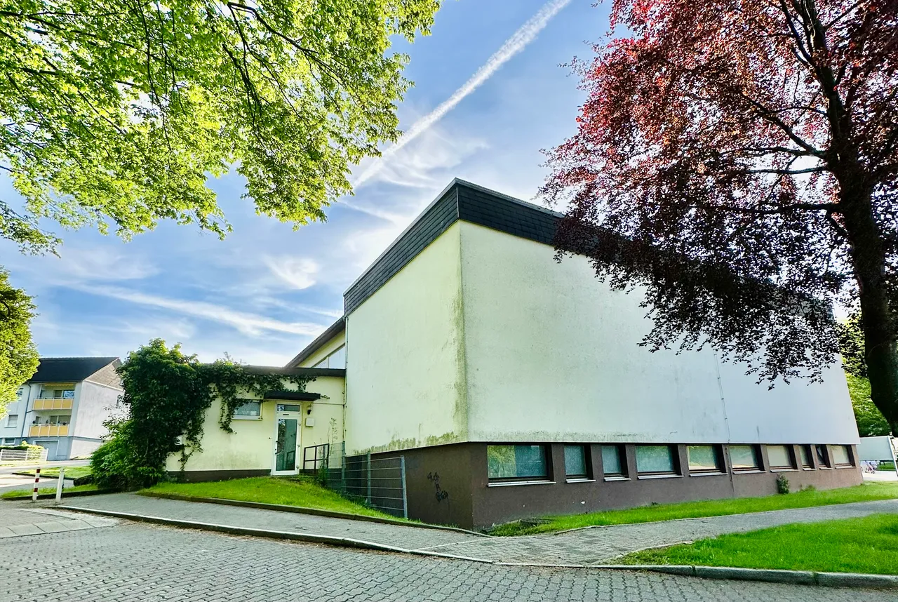 Helles, zweigeschossiges Gebäude mit dunklem Satteldach und einer Reihe von Fenstern, daneben ein Baum mit rotem Laub.