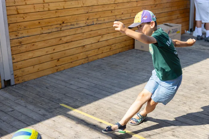Ein Junge in grünem T-Shirt und blauen Shorts tritt auf einer Holzterrasse gegen einen gelb-blauen Ball.