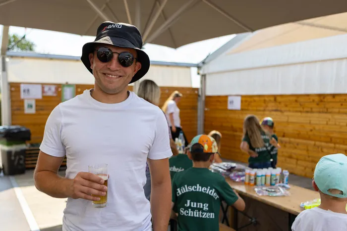 Ein lächelnder Mann mit Sonnenbrille und schwarzem Hut hält ein Glas Bier. Im Hintergrund sind Kinder in grünen T-Shirts bei einem Schützenfest zu sehen.