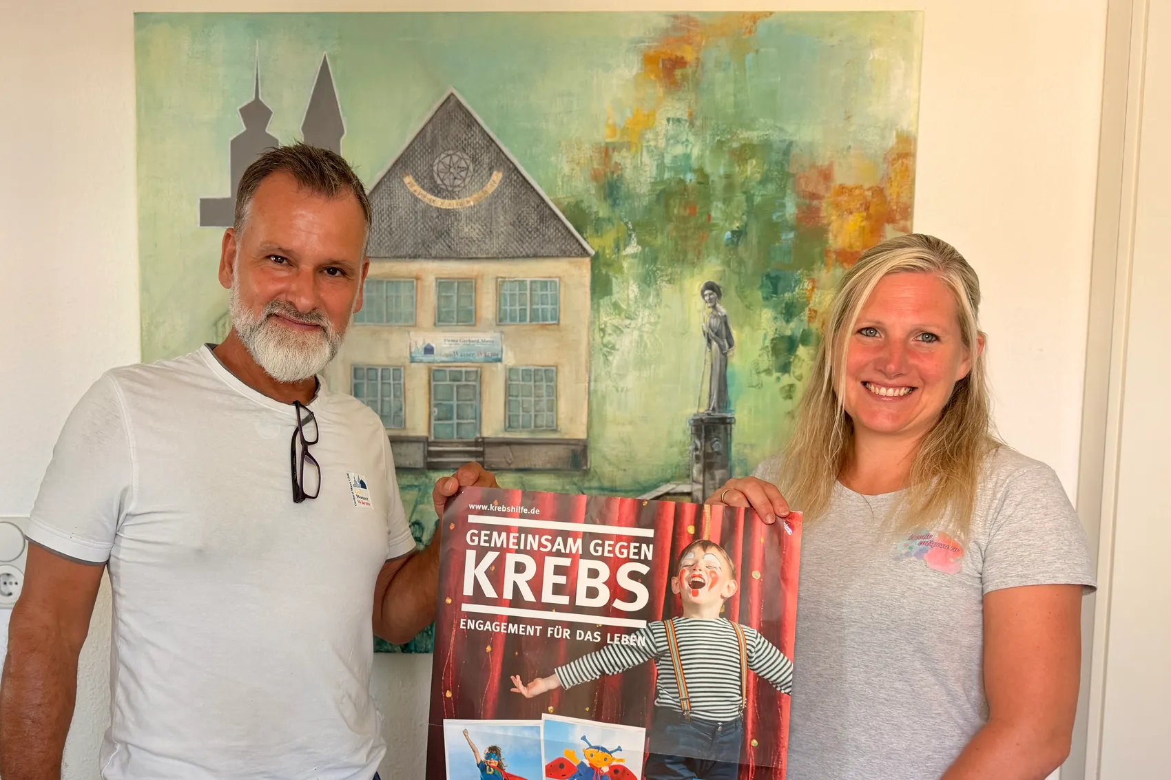 Ein Mann und eine Frau stehen vor einem Gemälde eines Hauses und halten ein Plakat mit der Aufschrift „Gemeinsam gegen Krebs“ und dem Logo der Deutschen Kinderkrebshilfe. Die Frau lächelt in die Kamera, während der Mann das Plakat hält.