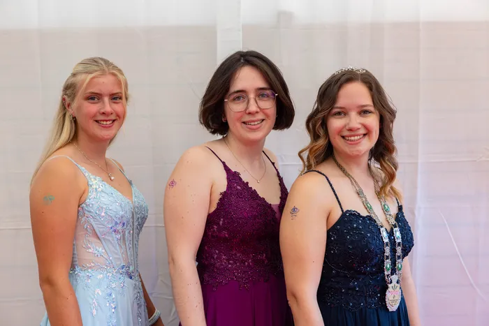 Drei junge Frauen in festlichen Kleidern in Hellblau, Lila und Dunkelblau lächeln vor einem weißen Vorhang.