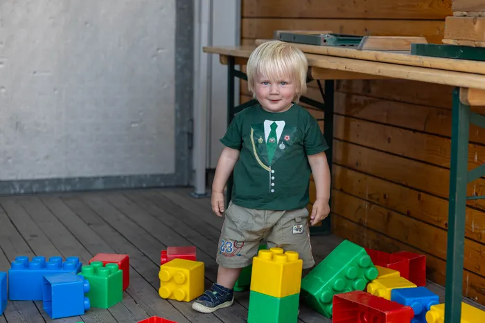 Ein kleiner Junge mit blonden Haaren steht zwischen großen, bunten Plastikbausteinen auf einem Holzboden.