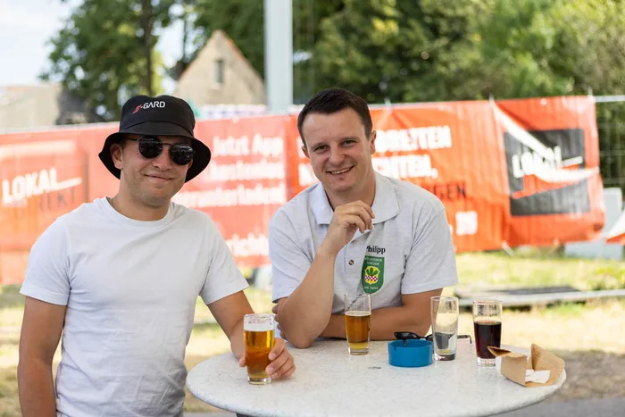 Zwei Männer stehen an einem runden Tisch mit Getränken vor einem orangefarbenen Banner im Freien.