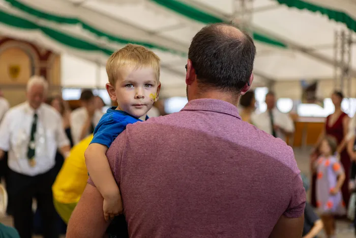 Ein kleiner Junge mit gelber Gesichtsbemalung wird von einem Mann auf dem Rücken getragen. Im Hintergrund sind Menschen in einem Festzelt zu sehen.
