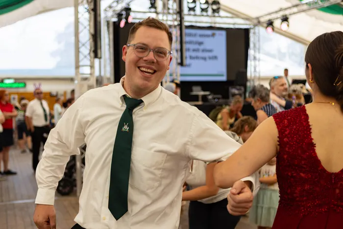 Ein lachender Mann in weißem Hemd und grüner Krawatte tanzt mit einer Frau in einem roten Kleid auf einem Schützenfest.