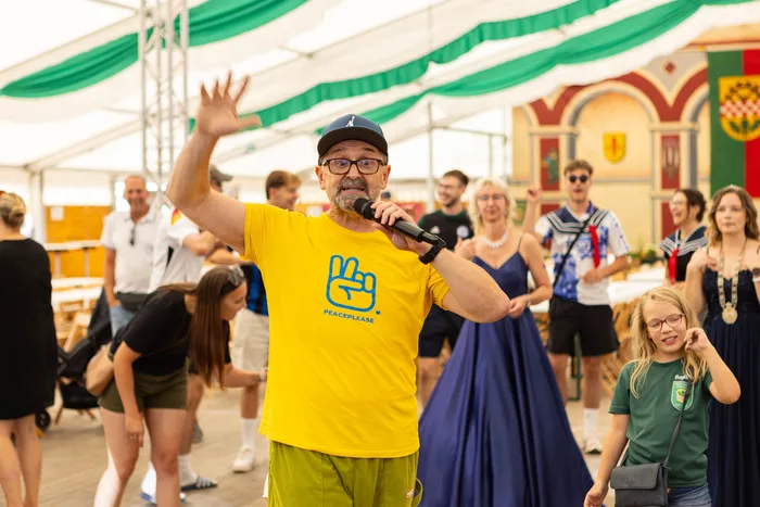 Ein Mann in einem gelben T-Shirt mit Peace-Logo spricht in ein Mikrofon und winkt bei einem Schützenfest.