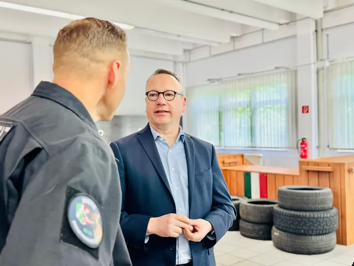 Ein Mann mit Brille in einem Blazer und Hemd spricht mit einem Mann in einer dunklen Jacke. Im Hintergrund sind Regale und Reifen zu sehen.