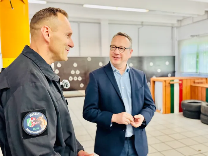 Ein Beamter in Uniform mit einem Abzeichen der Justiz und einem Mann in Anzug unterhalten sich in einem hellen Raum mit einer Schießwand im Hintergrund.