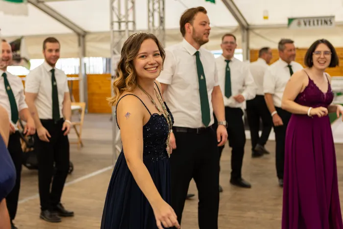 Eine lächelnde junge Frau in einem dunkelblauen Kleid steht in einem Festzelt, im Hintergrund stehen Männer in weißen Hemden und grünen Krawatten.