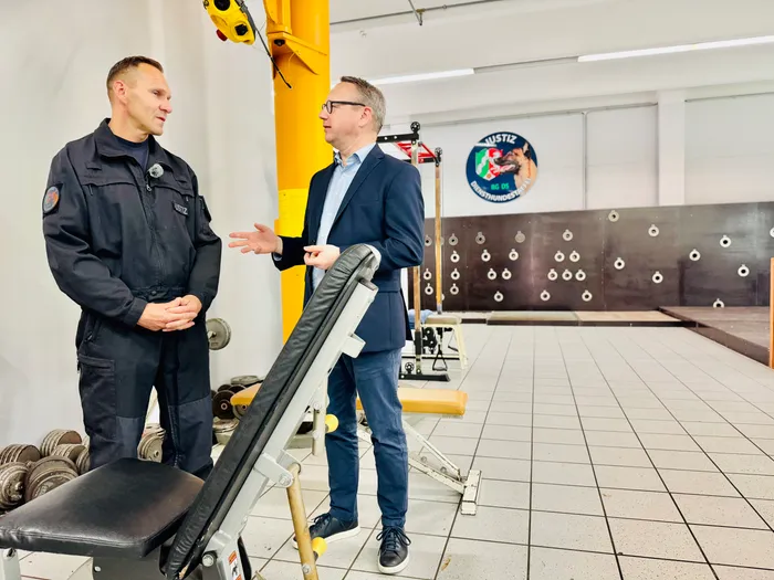 Zwei Männer stehen in einem Fitnessraum. Ein Mann in Uniform spricht mit einem Mann in Anzug, während er gestikuliert. Im Hintergrund ein Logo mit der Aufschrift 