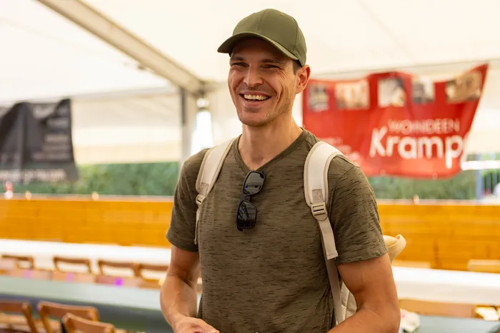 Ein lächelnder Mann mit grüner Kappe und Rucksack vor einem roten Banner unter einem Festzelt.