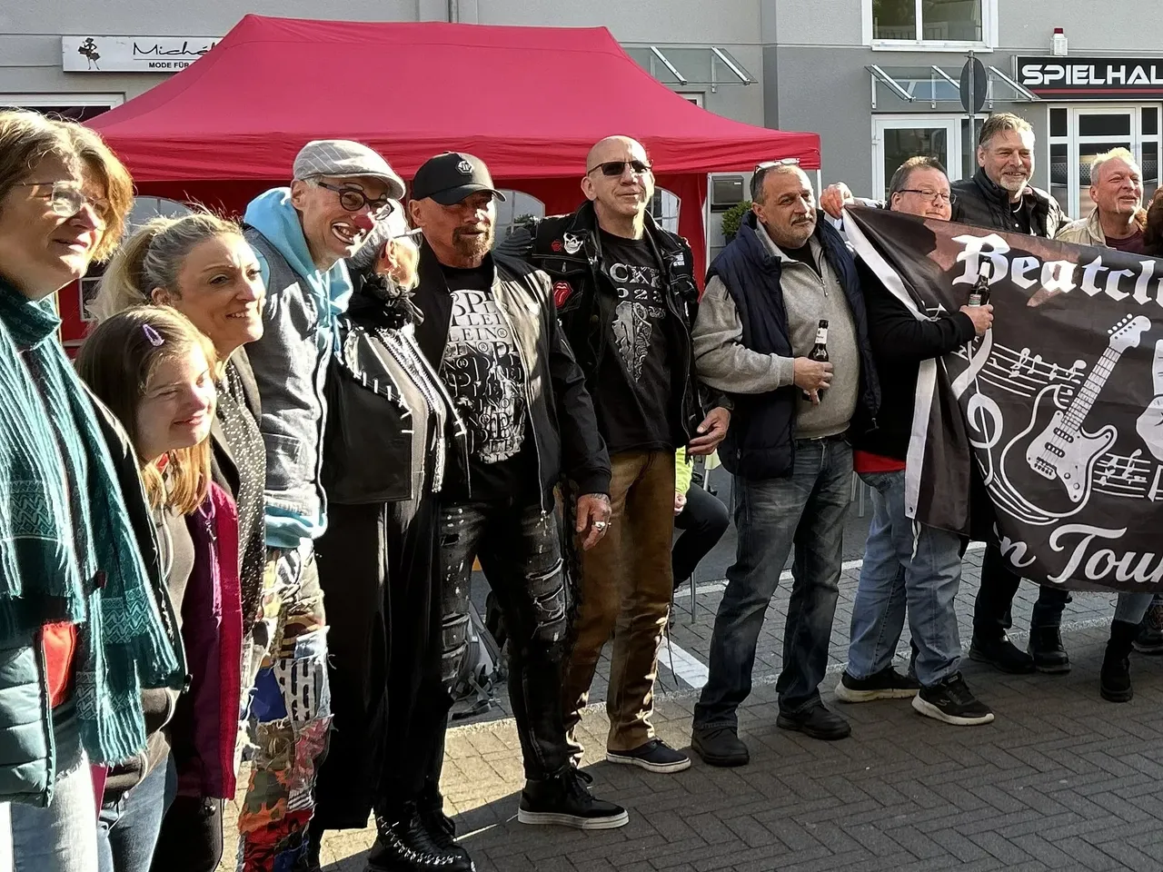 Eine Gruppe von Menschen steht vor einem roten Pavillon und einem Eiscafé, einige halten Getränke und ein Banner mit Gitarrenmotiven.