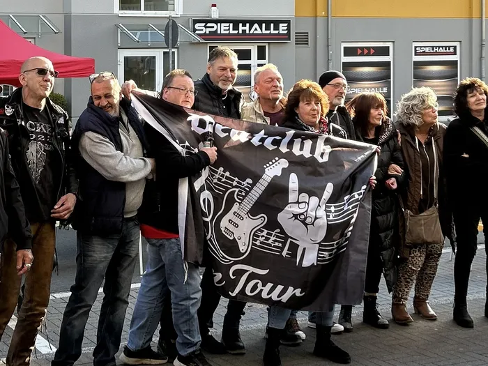 Eine Gruppe von Menschen hält ein Banner mit der Aufschrift „Beatclub“ und einer Gitarre sowie einer Hand mit dem „Rock’n’Roll“-Zeichen. Im Hintergrund ist eine Spielhalle zu sehen.