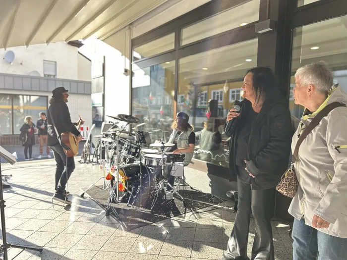 Eine Band spielt im Freien vor einem Eiscafé. Ein Sänger mit langem Haar hält ein Mikrofon, während eine Frau neben ihm zuhört.