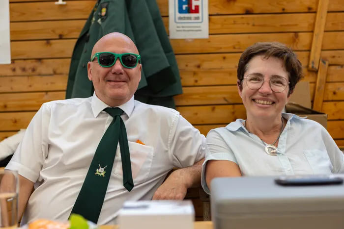 Ein lächelnder Mann mit grüner Sonnenbrille und grüner Krawatte sitzt neben einer lächelnden Frau mit Brille.