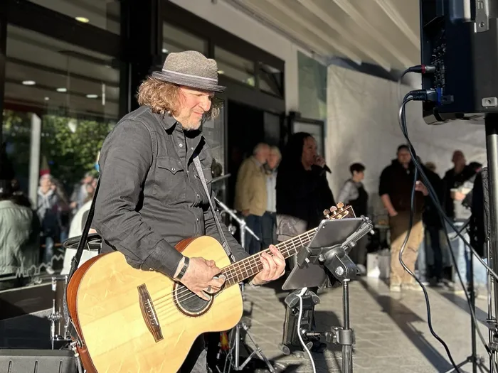 Ein Musiker mit Gitarre spielt vor einem Eiscafé im Freien, während Passanten vorbeigehen.