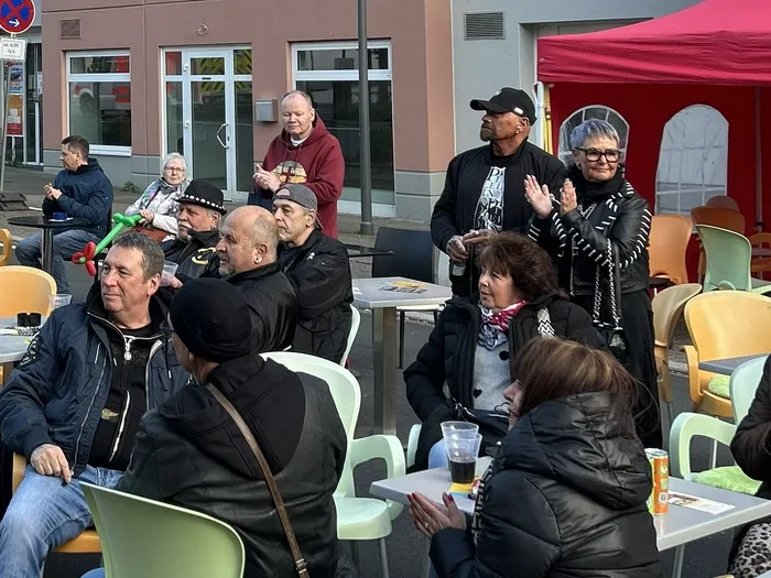 Zuschauer sitzen und stehen vor einem Eiscafé, einige klatschen. Im Hintergrund sind weitere Menschen und ein rotes Markisendach zu sehen.