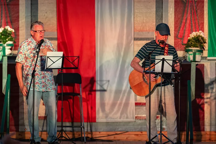 Ein Mann singt in ein Mikrofon, während ein anderer Mann auf einer Bühne Gitarre spielt, vor einem rot-weißen Banner.