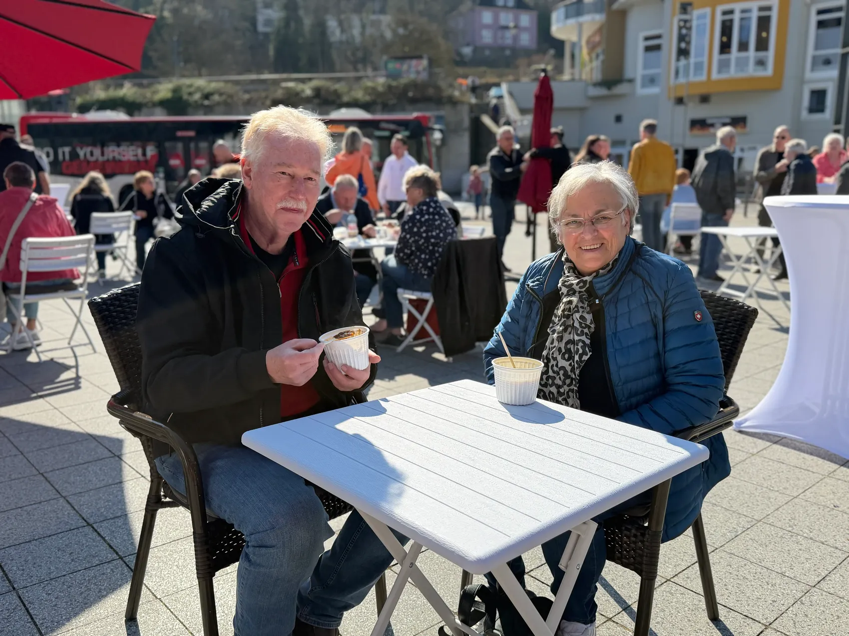 Zwei ältere Menschen sitzen an einem Tisch im Freien und halten jeweils eine Schale mit Speisen in der Hand, im Hintergrund weitere Besucher auf dem belebten Rathausplatz.
