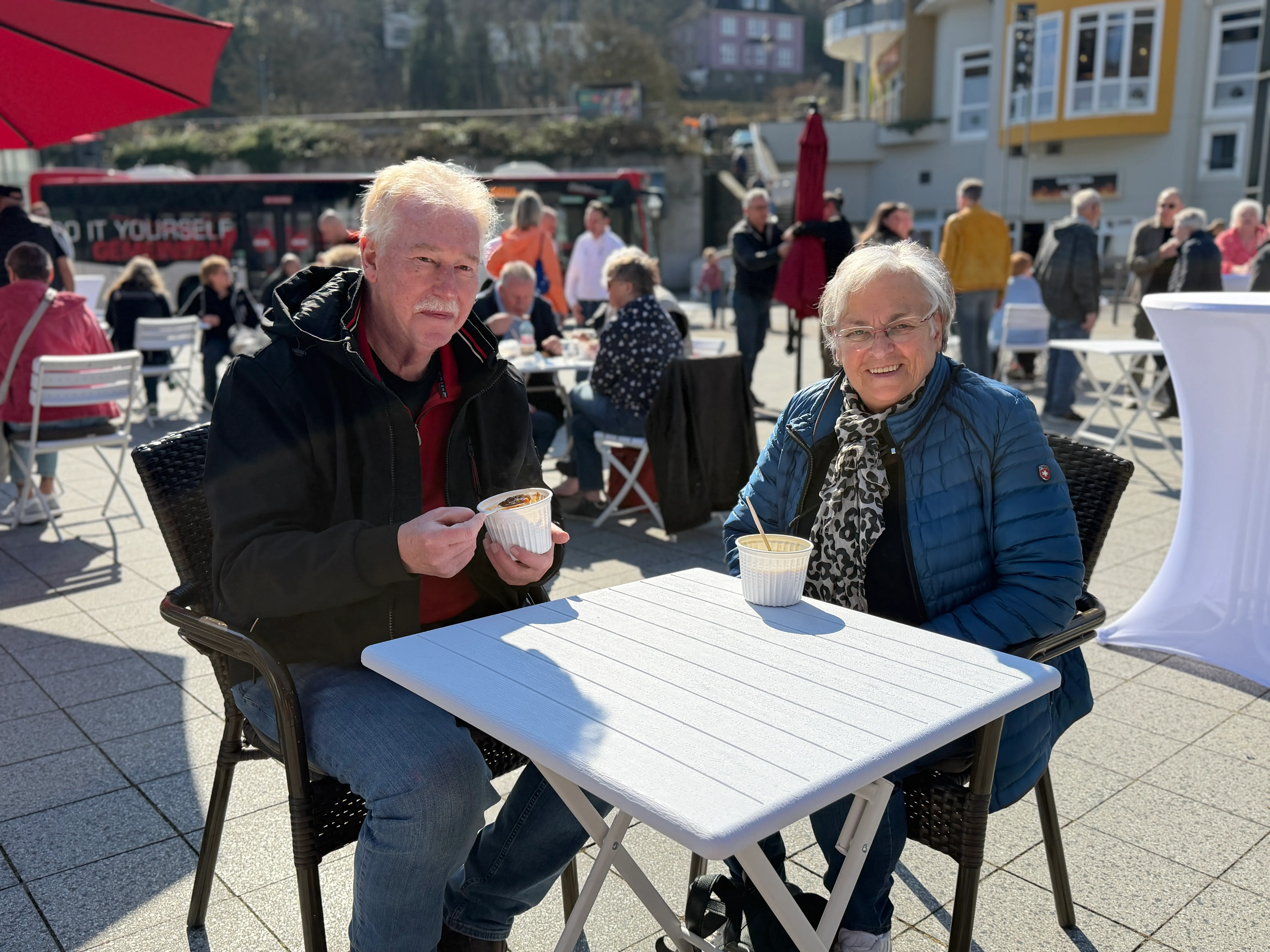 Zwei ältere Menschen sitzen an einem Tisch im Freien und halten jeweils eine Schale mit Speisen in der Hand, im Hintergrund weitere Besucher auf dem belebten Rathausplatz.