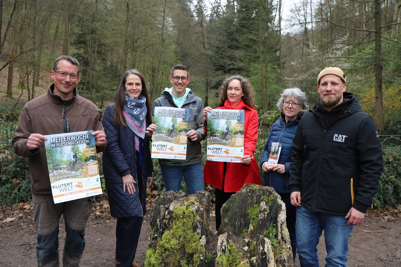 Sechs Personen stehen im Freien und halten jeweils ein farbiges Plakat mit dem Titel „Meilerwoche im Hülsenbecker Tal“ in der Hand.