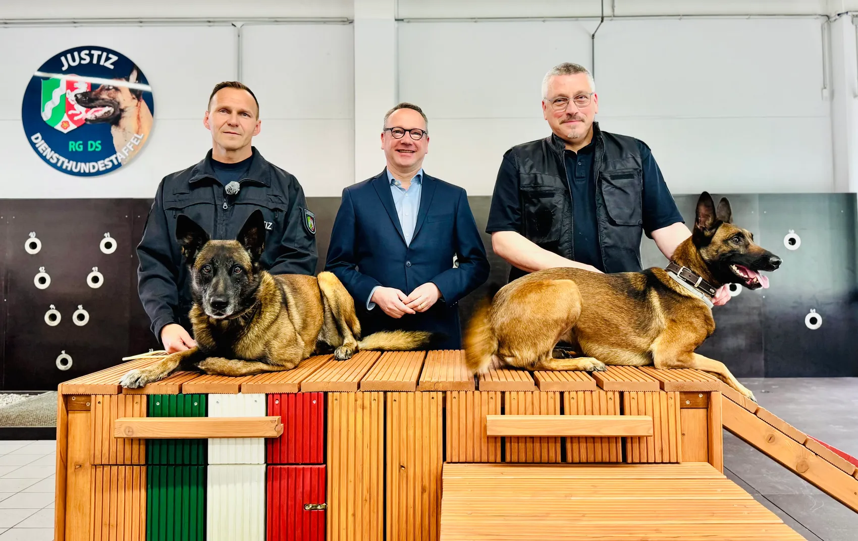 Drei Männer stehen mit zwei Deutschen Schäferhunden auf einer hölzernen Rampe. Links und rechts stehen Männer in Uniform, in der Mitte ein Mann im Anzug. Im Hintergrund ist ein Logo mit der Aufschrift 