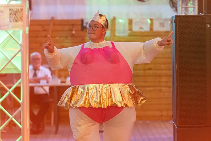 Eine Person in einem weiß-pinken Kostüm mit goldenem Rock posiert mit ausgestreckten Armen bei einem Schützenfest.