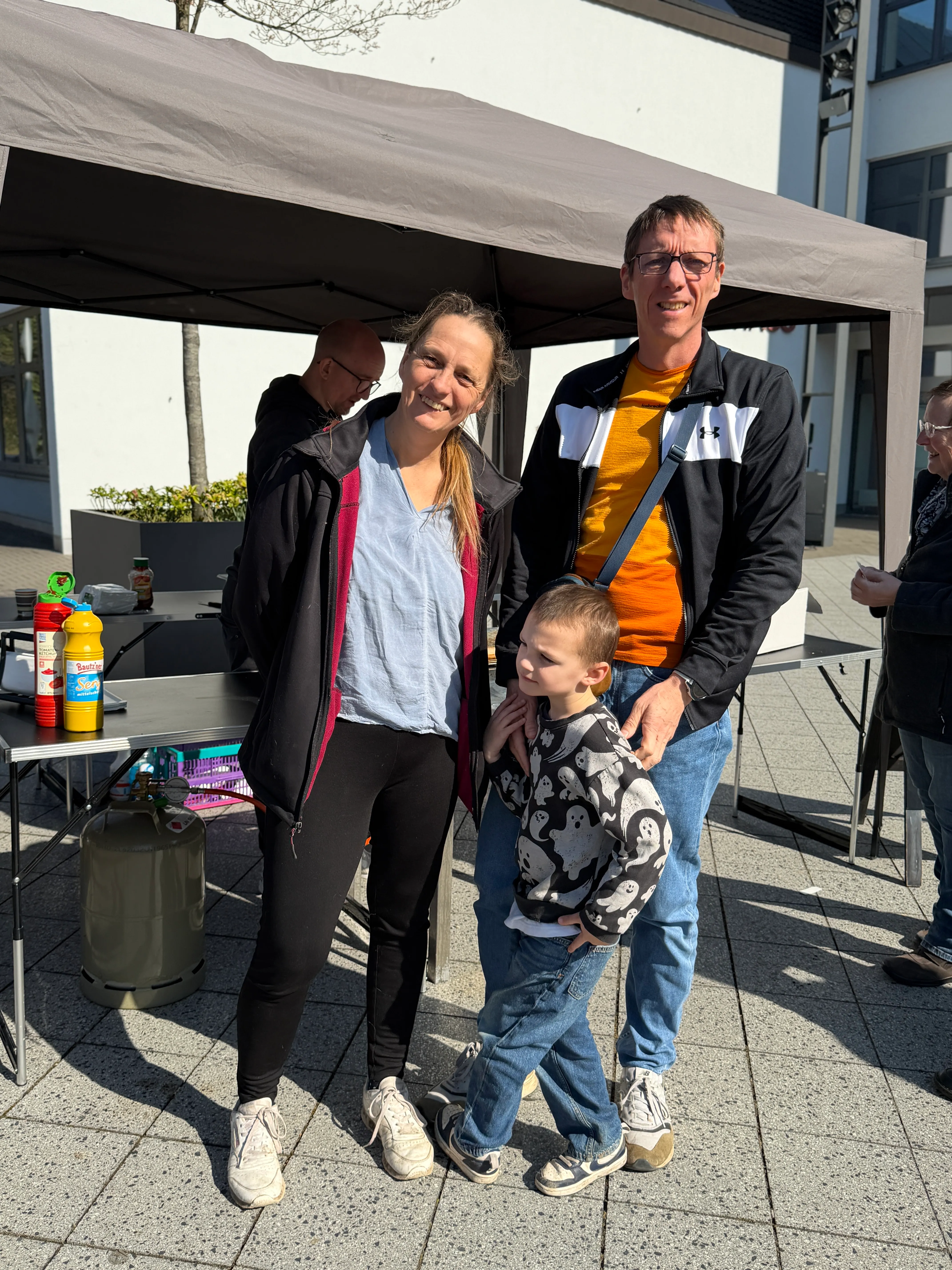 Drei Personen lächeln in die Kamera: eine Frau, ein Mann und ein kleines Kind. Im Hintergrund steht ein Pavillon und weitere Besucher sind zu sehen.