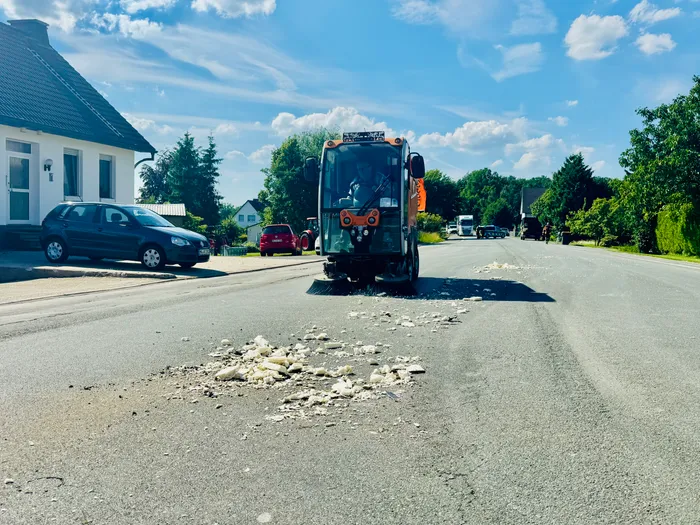 Ein orangefarbenes Straßenreinigungsfahrzeug steht auf einer Landstraße, umgeben von geparkten Autos und Häusern. Auf der Straße vor dem Fahrzeug liegt Schutt.