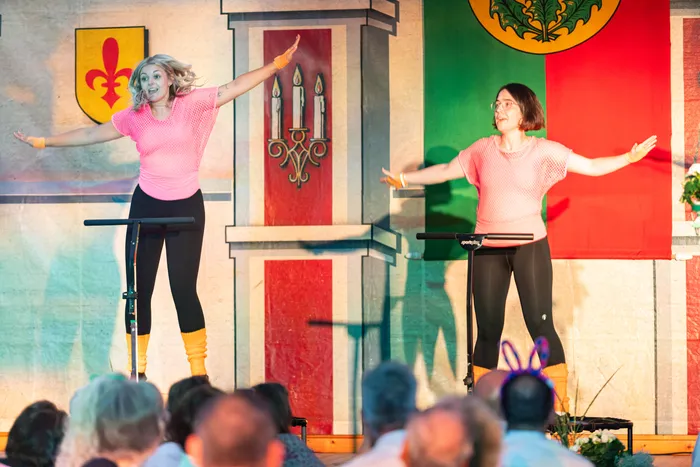 Zwei junge Performerinnen in rosa Oberteilen und schwarzen Leggings tanzen mit ausgebreiteten Armen auf einer bunt gestalteten Bühne.