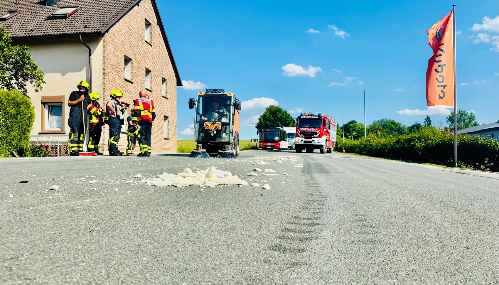 Feuerwehrleute stehen neben Trümmern auf der Straße, während ein Räumfahrzeug und ein Feuerwehrauto im Hintergrund zu sehen sind. Ein rotes Banner weht am Straßenrand.