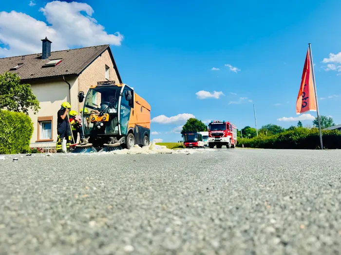 Feuerwehrleute sind mit Aufräumarbeiten beschäftigt, nachdem ein Fahrzeug in ein Gebäude gefahren ist. Ein rotes Feuerwehrauto steht in der Nähe, während Trümmer auf der Straße liegen.