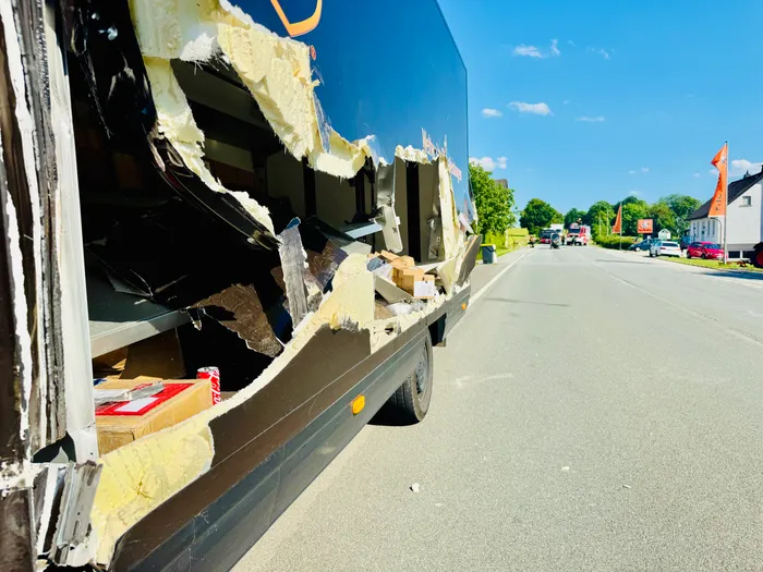 Der hintere Teil eines beschädigten Lastwagens ist zu sehen, wobei Isolierung und Kartons aus einer großen Öffnung im Inneren ragen. Im Hintergrund ist eine Landstraße mit Gebäuden und anderen Fahrzeugen zu erkennen.