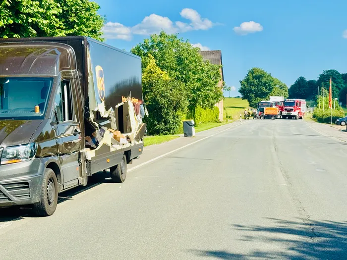Ein beschädigter UPS-Lieferwagen steht am Straßenrand, während im Hintergrund Einsatzfahrzeuge der Feuerwehr und Polizei zu sehen sind. Der Unfallort befindet sich auf einer Landstraße mit Bäumen und Feldern.