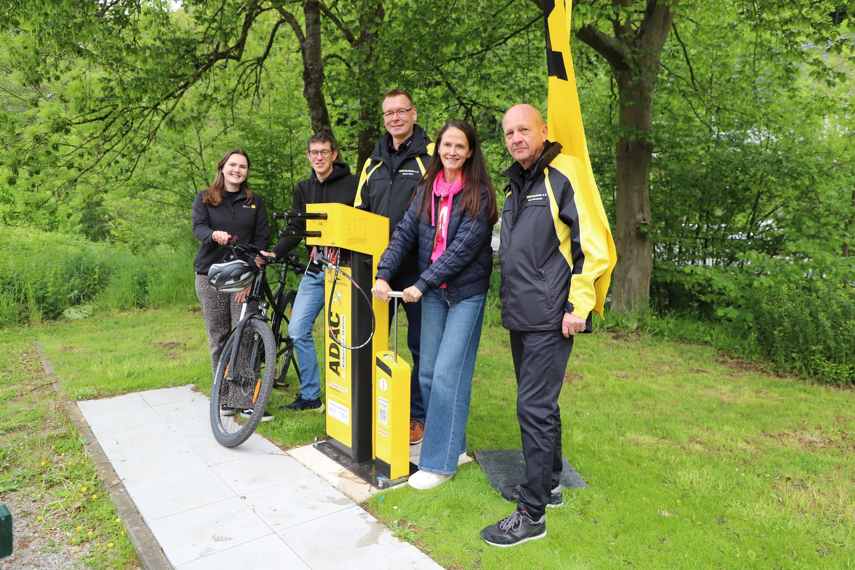 Vier Personen stehen vor einer gelben Radservicestation mit Werkzeug und Luftpumpe, neben einem Fahrrad. Im Hintergrund sind Bäume und Grünflächen zu sehen.