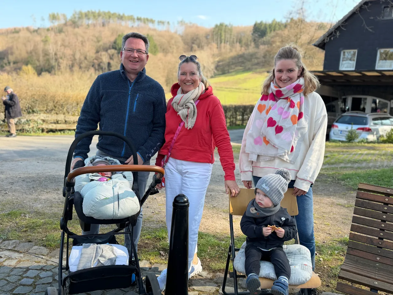 Familie mit Kinderwagen und kleinem Kind vor einem Bauernhaus an einem sonnigen Tag. Im Hintergrund eine grüne Landschaft und parkende Autos.