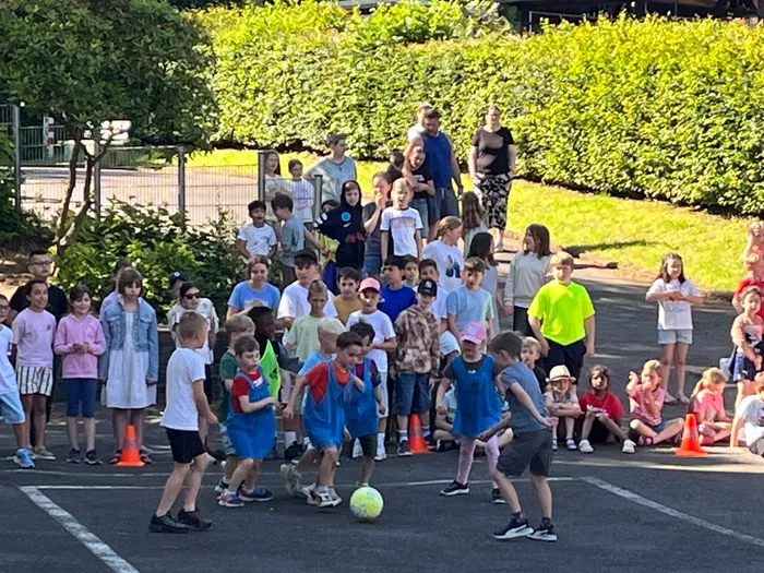 Eine Gruppe von Kindern spielt Fußball auf einem Parkplatz, umgeben von Zuschauern und Absperrhütchen. Im Hintergrund stehen Erwachsene und weitere Kinder auf einer Anhöhe.