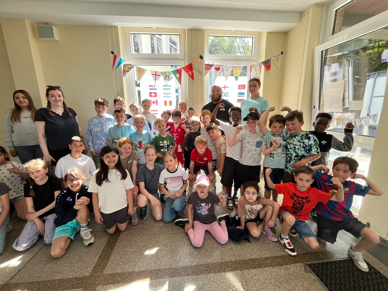 Eine Gruppe von Kindern und einigen Erwachsenen posiert für ein Foto. Viele der Kinder zeigen ihre Muskeln. Im Hintergrund sind Flaggen verschiedener Länder zu sehen.