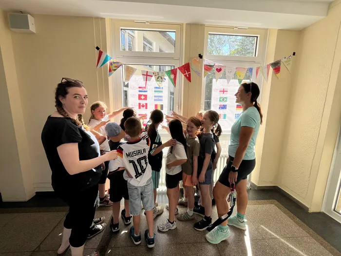 Eine Gruppe von Kindern steht mit zwei Betreuerinnen vor einem Fenster, das mit Flaggen der teilnehmenden Nationen der Fußball-Weltmeisterschaft dekoriert ist. Einige Kinder zeigen auf die Flaggen.