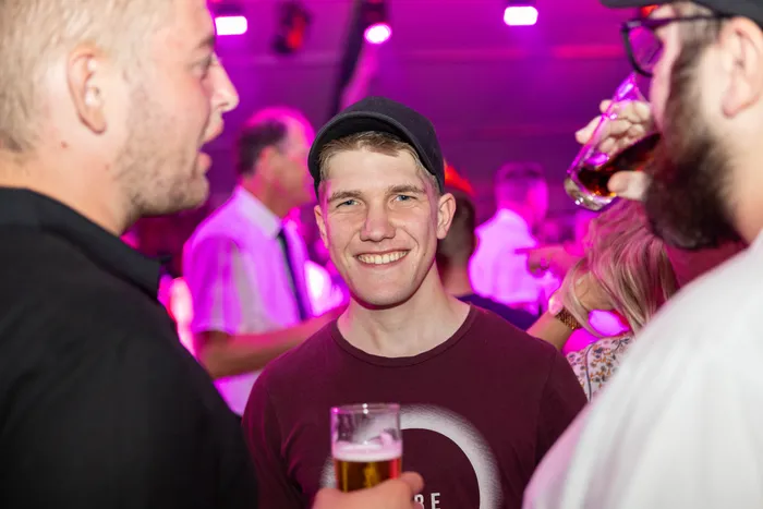 Ein lächelnder junger Mann mit schwarzer Kappe hält ein Glas Bier bei einer pink beleuchteten Abendveranstaltung.