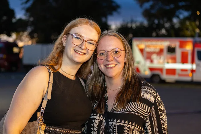 Zwei lächelnde junge Frauen mit Brillen stehen eng beieinander vor einem unscharfen Rettungswagen bei Nacht.