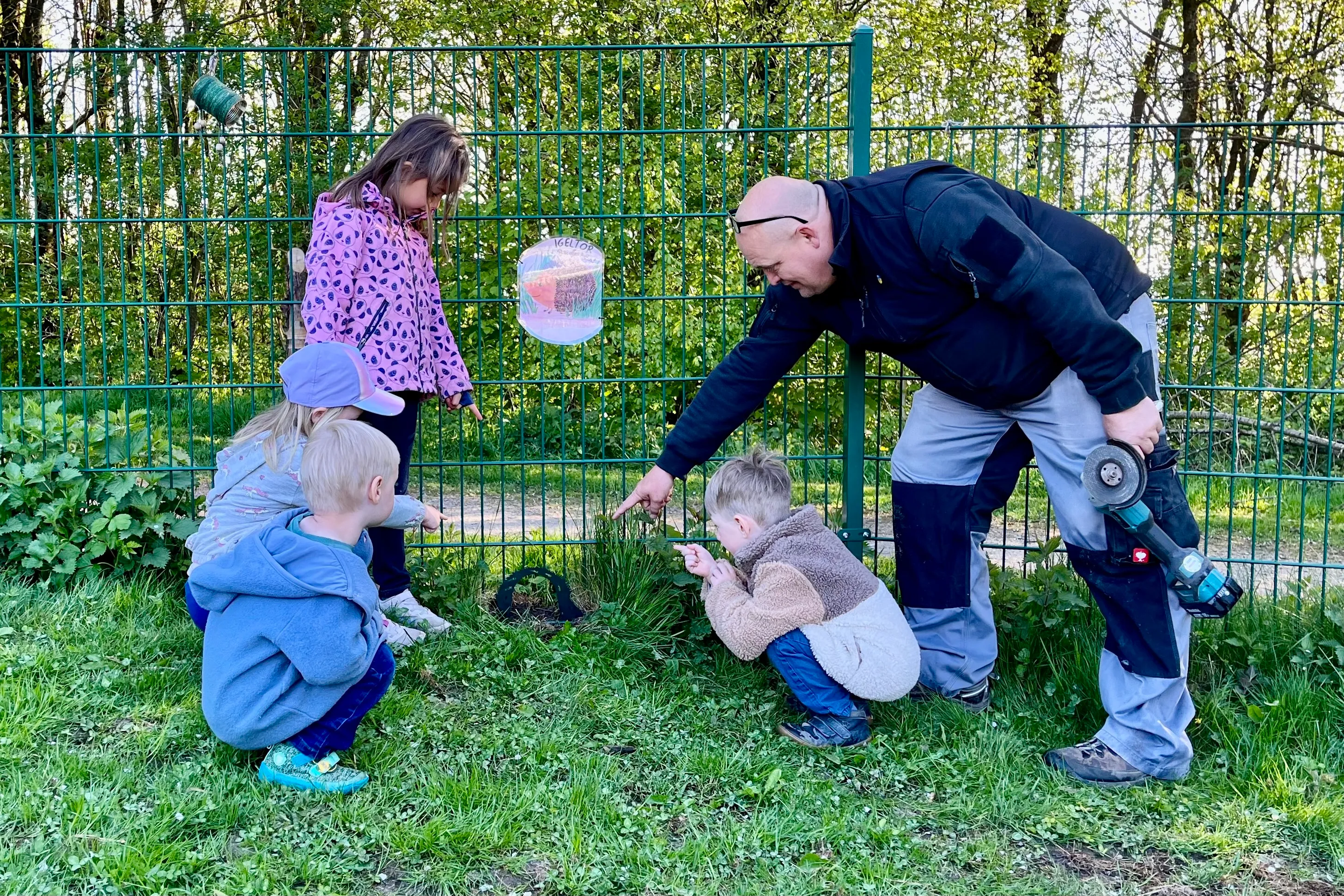 Ein Mann zeigt zwei Kindern auf dem Rasen einen Bereich unter einem Zaun, während ein weiteres Kind im Hintergrund steht. Im Hintergrund ist ein Tor mit einem Schild zu sehen.