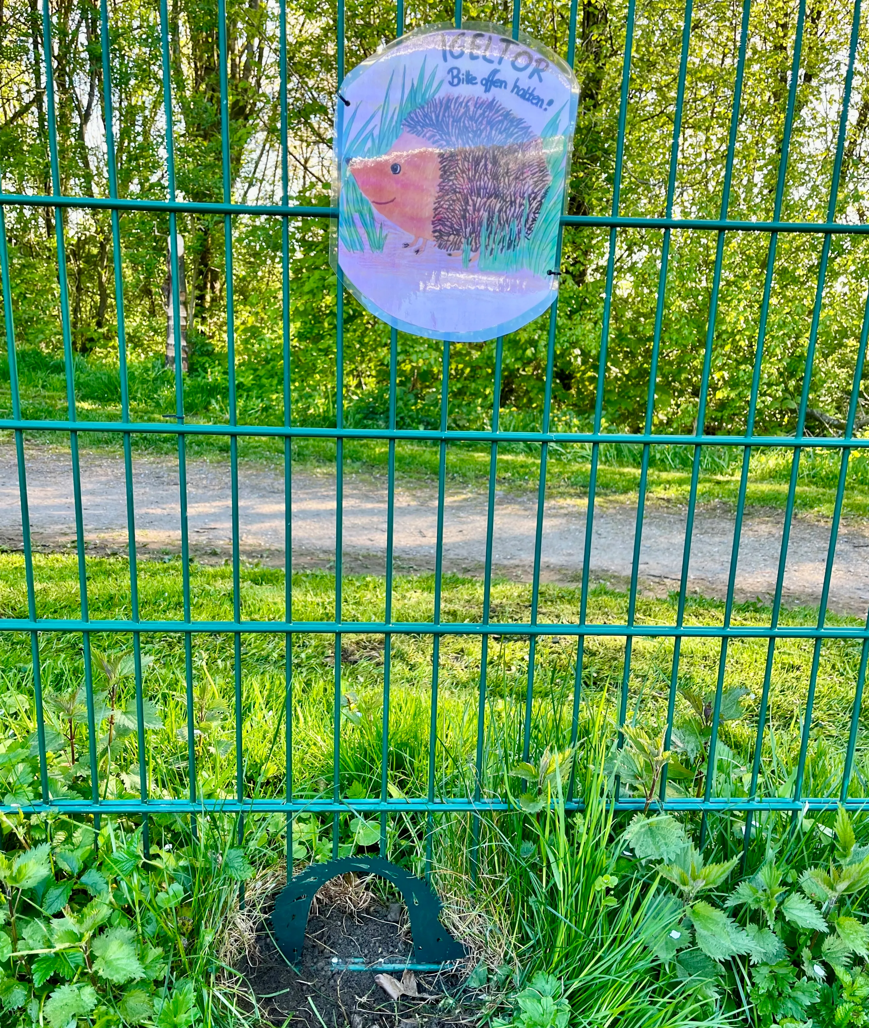 Ein Schild mit einem Igel an einem grünen Zaun, beschriftet mit „Igeltor – Bitte offen halten!“. Im Hintergrund ist ein Weg zu sehen.