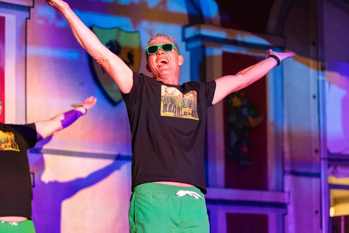 Ein Mann mit grünen Sonnenbrillen und weit ausgebreiteten Armen jubelt auf einer bunt beleuchteten Bühne beim Schützenfest.