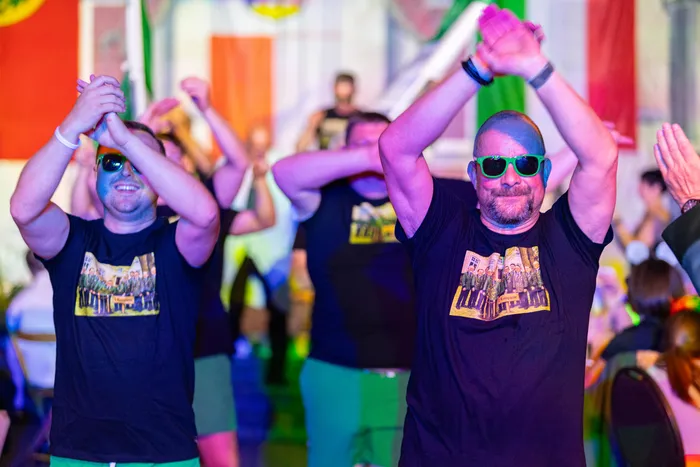 Männer in schwarzen T-Shirts mit Sonnenbrillen tanzen mit erhobenen Armen bei einem bunten Dorfabend während des Schützenfests.