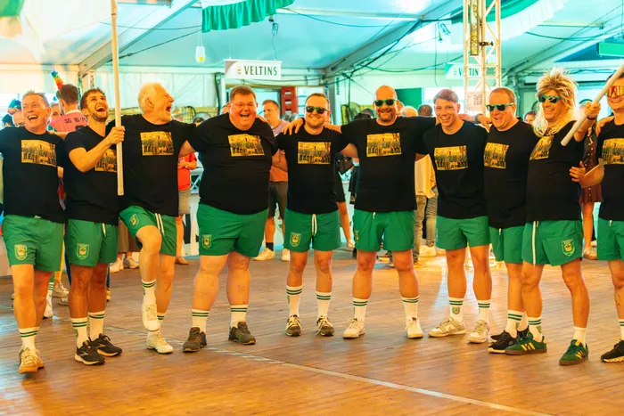 Eine Gruppe lachender Männer in einheitlichen schwarzen T-Shirts und grünen Shorts posiert gemeinsam in einem Festzelt.